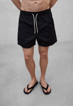 Mann trägt schwarze Badeshorts mit weißen Kordeln und schwarze Flip-Flops, steht vor einem schlichten grauen Hintergrund.