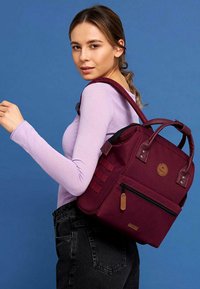 Sac à dos bordeaux en toile avec deux poignées supérieures et des sangles réglables. Il comprend une poche zippée à l'avant et un patch logo.