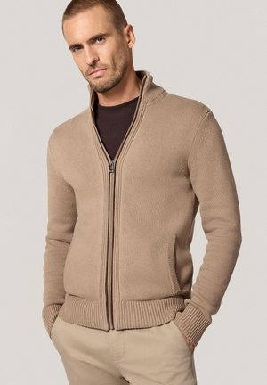 Beżowy sweter na zamek z prążkowanymi mankietami i dołem, z teksturowanym wzorem dzianiny i wysokim kołnierzem. Noszony na ciemną bluzkę.