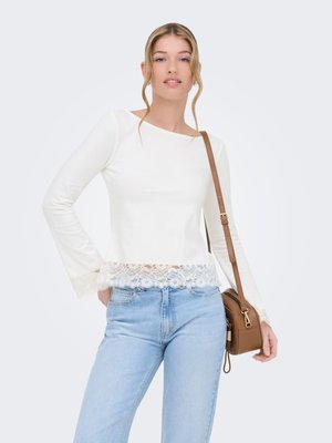 Young woman wearing a long-sleeve white top with lace trim, light blue jeans, and carrying a brown shoulder bag on a white background.
