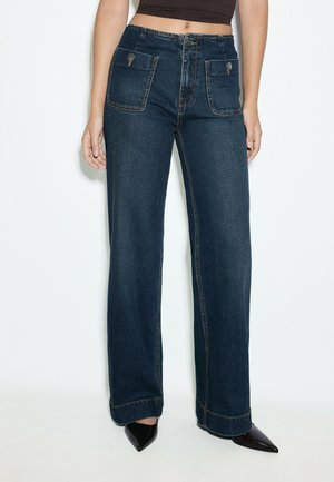Femme portant un jean large bleu foncé avec des poches plaquées à l'avant et des chaussures noires à bout pointu sur fond blanc.