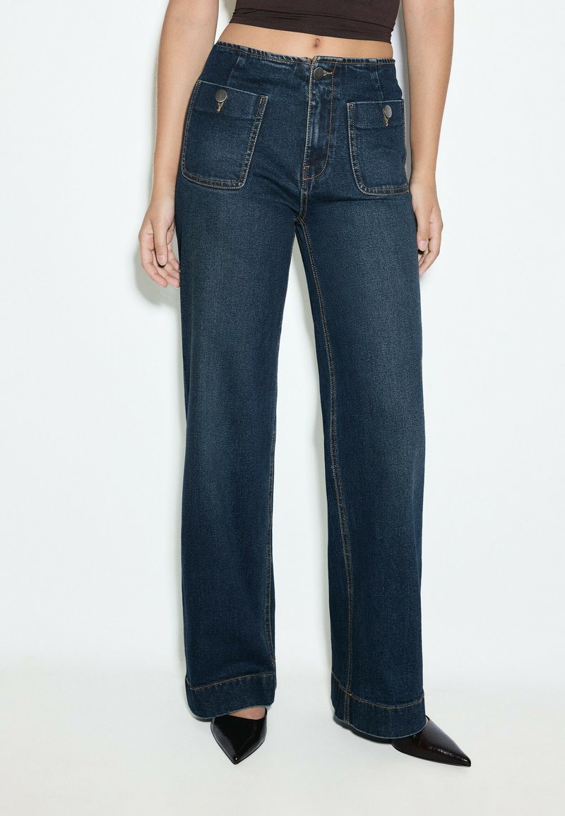 Femme portant un jean large bleu foncé avec des poches plaquées à l'avant et des chaussures noires à bout pointu sur fond blanc.