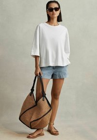 Hvid oversized t-shirt med korte ærmer, denimshorts og brune flade sandaler. Holder en stor vævet stråtaske med sort kant.