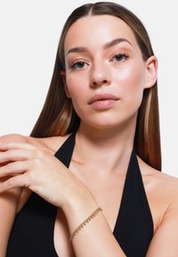 Bracelet en or avec des accents pendants, présentant un design de chaîne délicat. Une modèle porte un haut asymétrique noir, mettant en valeur le bracelet sur un poignet.