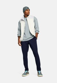 Chaleco de forro polar blanco sobre un sudadera azul clara, pantalones marinos y zapatillas gris claro con acentos azules. Un gorro de lana gris completa el look.