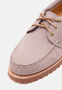 Nærbillede af taupefarvet lædersko med beige syning, brune ruskindssnørebånd, metaløjer og tyk, lysebrun gummisål med dybe mønstre.