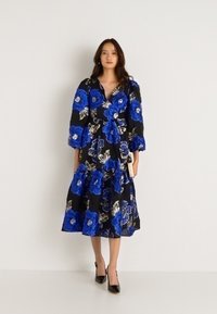 Λουλουδάτο midi φόρεμα με μαύρη βάση, διακοσμημένο με μεγάλες μπλε ανθοσύνθεσεις. Μακριά μανίκια, βαθύ V-ντεκολτέ και ροή σε φούστα με στρώσεις. Μαύρες γόβες.