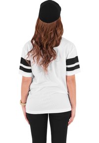 Fehér hálós mez fekete csíkokkal az ujjakon, fekete sapkával párosítva. Hosszú haj hullámzik a háton, slim-fit fekete nadrággal viselve.