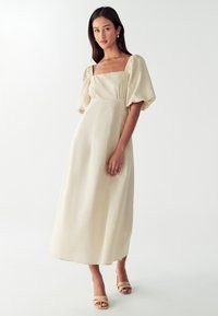 Kremasta midi haljina s nabranim rukavima, četvrtastim dekolteom i glatkom teksturom. Uparena s bež sandalama na blok petu.