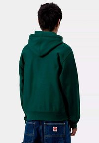 Sudadera verde de corte holgado, con una capucha grande y puños acanalados. Combinada con denim azul, que muestra un diseño de bolsillo.
