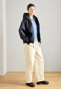 Gepolsterte schwarze Kapuzenjacke mit Seitentaschen, kombiniert mit einem hellblauen Hemd mit Knopfleiste und beigen weiten Hosen. Schwarze Plateauschuhe.