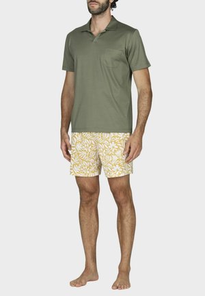 Uomo che indossa una polo verde oliva a maniche corte e pantaloncini gialli con motivo floreale, in piedi a piedi nudi su uno sfondo uniforme.