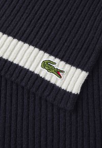 Maglione blu navy a coste con un bordo spesso a righe bianche. Presenta un logo di un coccodrillo verde ricamato con accenti rossi sul lato.