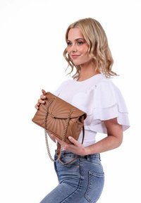 Hnědá prošívaná kabelka s řetízkovým popruhem; má klopu a dekorativní kování. Model ji drží proti jednobarevnému pozadí.