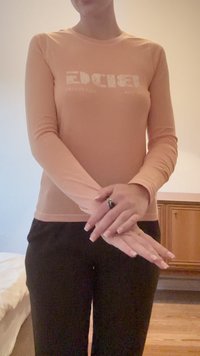 Camicia a maniche lunghe di colore arancione chiaro con design in testo bianco, abbinata a pantaloni neri. Il tessuto appare liscio e aderente, con scollo rotondo.