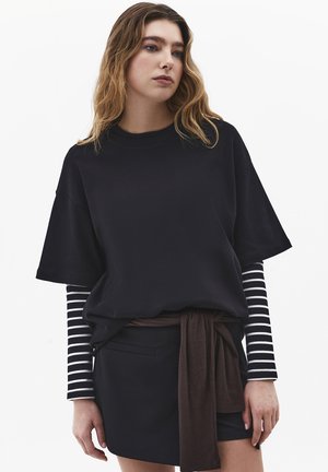 Młoda kobieta o falowanych włosach, ubrana w czarny oversize T-shirt na czarno-białym paskowanym topie z długim rękawem, czarną spódnicę i brązowy związany sweter.