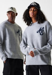 Lys grå strikkede gensere med en myk tekstur, med en mørk blå logo på brystet. Modellen har på seg en beigefarget caps, og en annen har på seg en svart caps.
