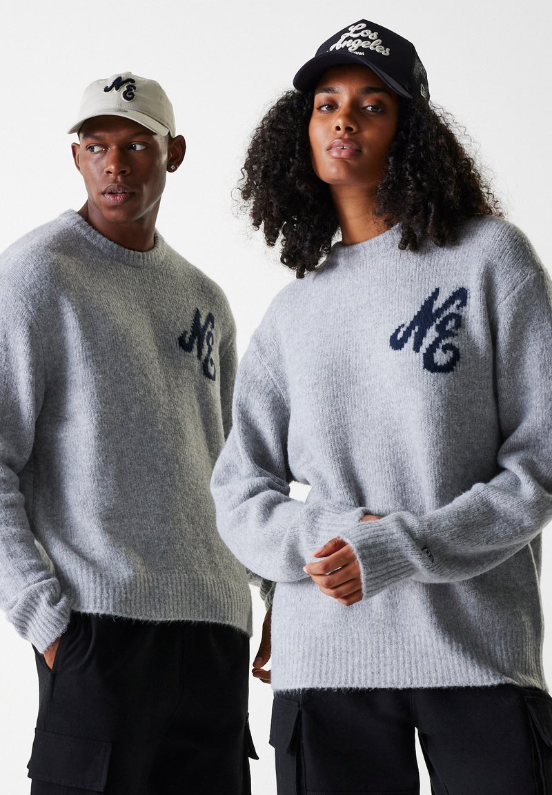 Lys grå strikkede gensere med en myk tekstur, med en mørk blå logo på brystet. Modellen har på seg en beigefarget caps, og en annen har på seg en svart caps.