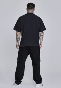 Melna, pārāk liela t-kreklā ar īsām piedurknēm, apvienota ar melniem kravas biksēm ar sānu kabatām, attēlota uz neitrāla fona.