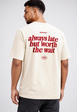 Man with short curly hair wearing a cream t-shirt with red text "always late but worth the wait" standing against a plain background.