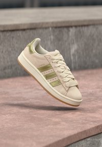 Μπεζ σουέντ αθλητικό παπούτσι με πράσινες λεπτομέρειες, τρεις πλευρικές ρίγες και λαστιχένια σόλα. Διαθέτει στρογγυλή μύτη και σχέδιο με κορδόνια.