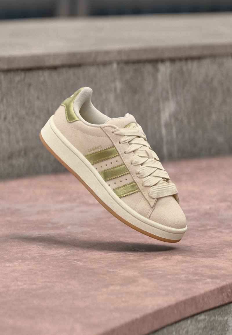 Ténis de camurça bege com detalhes verdes, três riscas laterais e sola de borracha. Apresenta biqueira arredondada e design com atacadores.