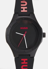 Black watch with a matte finish, round face, red accents, white hour and minute hands, and silicone strap printed with "HUGO."