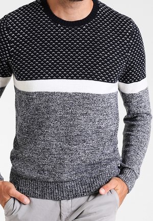 Homme portant un pull en maille à blocs de couleurs bleu marine, blanc et gris avec une partie supérieure à motifs et une partie inférieure unie, les mains dans les poches d’un pantalon gris clair.