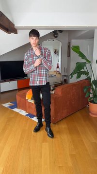 Νεαρός άνδρας που ρυθμίζει το ρολόι του, φορώντας καρό πουκάμισο και μαύρο παντελόνι, στέκεται σε ένα μοντέρνο σαλόνι με πορτοκαλί καναπέ, τηλεόραση και μεγάλο πράσινο φυτό.