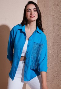Camicia blu a trama con maniche lunghe, chiusura con bottoni e dettaglio di pizzo nella tasca. Abbinata a pantaloni bianchi a vita alta.