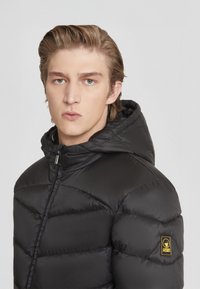Giacca a vento nera con cappuccio, caratterizzata da un design trapuntato, zip frontale e un patch con logo giallo sul braccio sinistro. Texture liscia.