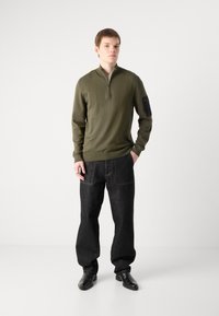 Dunkelgrüner Reißverschluss-Pullover mit gerippten Bündchen und einem schwarzen Ärmel-Patch, kombiniert mit weit geschnittenen, dunkelblauen Jeans und schwarzen Schuhen.