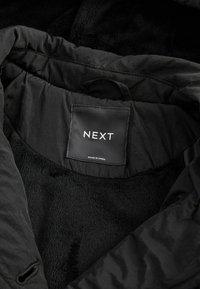 Veste noire avec doublure douce affichant une étiquette noire à l'intérieur portant les mentions "NEXT" et "Made in China" sous la zone du col.