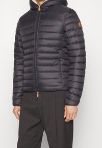 Giubbotto puffer nero con trapuntatura orizzontale, zip frontale e cappuccio. Caratterizzato da un tirazip arancione e logo sulla manica sinistra. Materiale testurizzato.