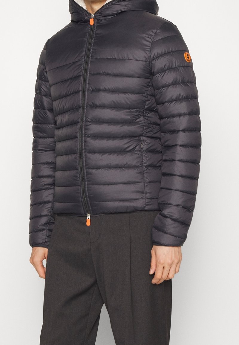 Giubbotto puffer nero con trapuntatura orizzontale, zip frontale e cappuccio. Caratterizzato da un tirazip arancione e logo sulla manica sinistra. Materiale testurizzato.