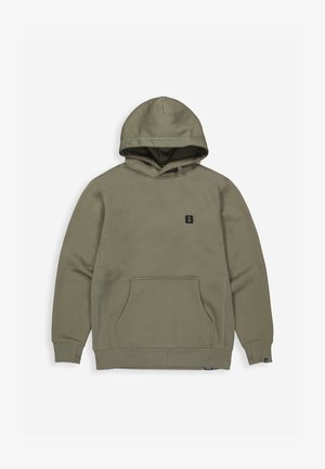 Man die een olijfgroene capuchontrui en bijpassende joggingbroek draagt, staand tegen een effen lichte achtergrond, naar voren gericht met een neutrale uitdrukking.