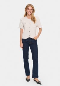 Lichtbeige, kortemouwen overhemd met knopen en twee borstzakken, gecombineerd met donkerblauwe rechte jeans en zwarte puntige schoenen.