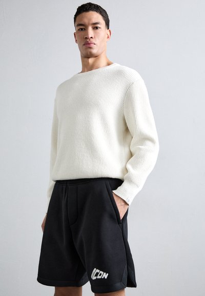 Pull en tricot crème à côtes associé à un short de sport noir avec un logo blanc. Le pull a un décolleté large et des manches longues.