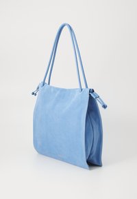 Sac fourre-tout en suède bleu avec deux longues anses, texture lisse, forme douce, et un détail logo subtil près du bas. Présente un design simple.
