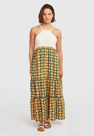 Ženska stoji oblečena v brezrokavno belo pleteno obleko s krilom oranžnih in kremnih cvetličnih vzorcev ter belimi sandali.