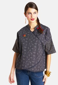 Dunkelbluse mit kurzen Ärmeln und einem weißen Muster, roten Akzenten und goldenen Beschlägen; kombiniert mit blauen Jeans und Armreifen.