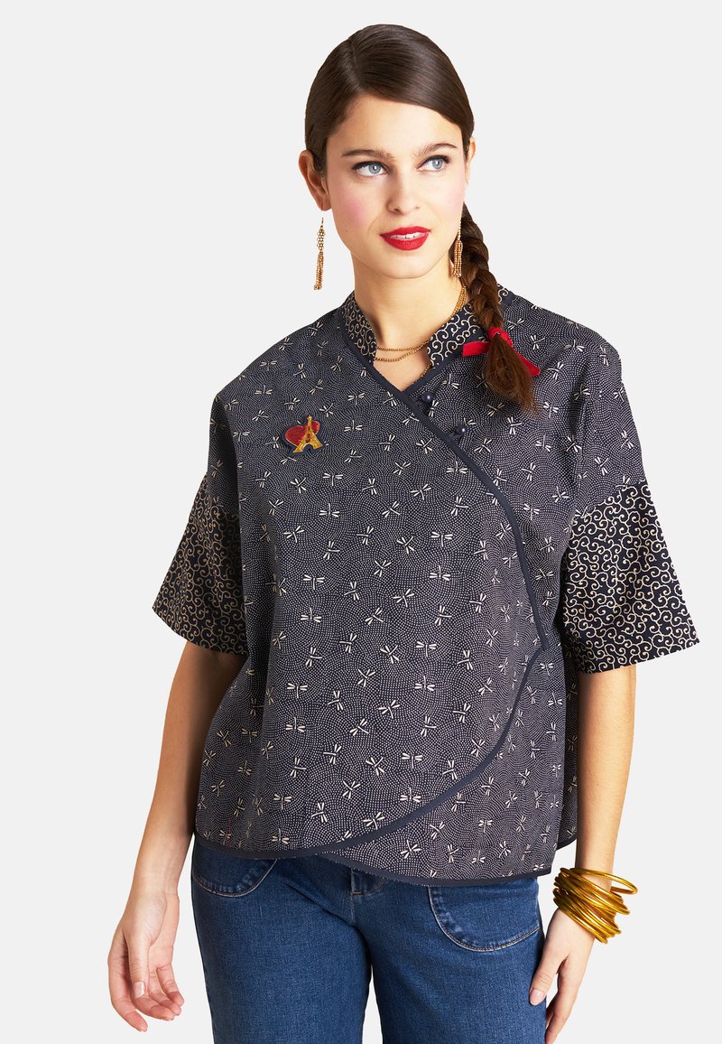 Dunkelbluse mit kurzen Ärmeln und einem weißen Muster, roten Akzenten und goldenen Beschlägen; kombiniert mit blauen Jeans und Armreifen.