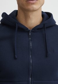 Felpa navale con zip realizzata in tessuto spesso, con cappuccio regolabile, chiusura a zip e logo discreto sul lato sinistro del petto.