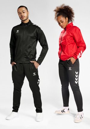 Zwarte sportieve jas en broek met wit logo en chevrondesigns. Rode ritsjas gecombineerd met zwarte broek. Beide modellen dragen sportief schoeisel.