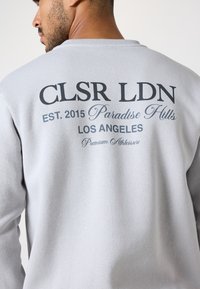 Grauer Sweatshirt mit lockerer Passform, Rundhalsausschnitt und großen schwarzen Text auf der Rückseite, einschließlich Markenname und Standortdetails.