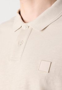 Polo beige avec un col côtelé et deux boutons. Sur le coin supérieur gauche, il y a un patch texte de la marque en relief. Texture de tissu douce.