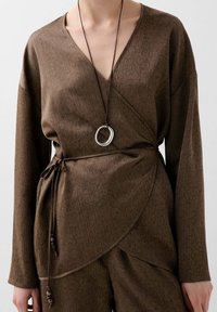 Top de envoltura texturizada marrón con escote en V, mangas largas y un lazo en la cintura. Presenta un collar con un colgante plateado redondeado.