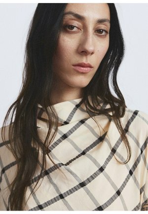 Donna con capelli lunghi e scuri, indossa una maglia beige a collo alto con motivo a quadri nero e grigio, espressione neutra, sfondo chiaro.