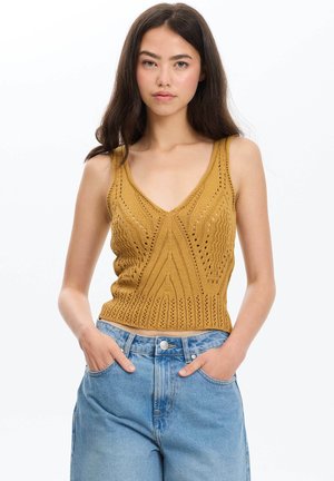Joven mujer con cabello largo y oscuro, vistiendo una camiseta sin mangas de punto color mostaza y vaqueros de talle alto en azul claro con las manos en los bolsillos.