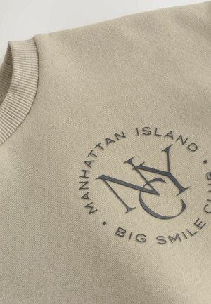 Beige Sweatshirt mit geripptem Ausschnitt, ausgestattet mit einem dunkelgrauen runden Logo, auf dem "MANHATTAN ISLAND" und "BIG SMILE CLUB" steht. Weiches Material.